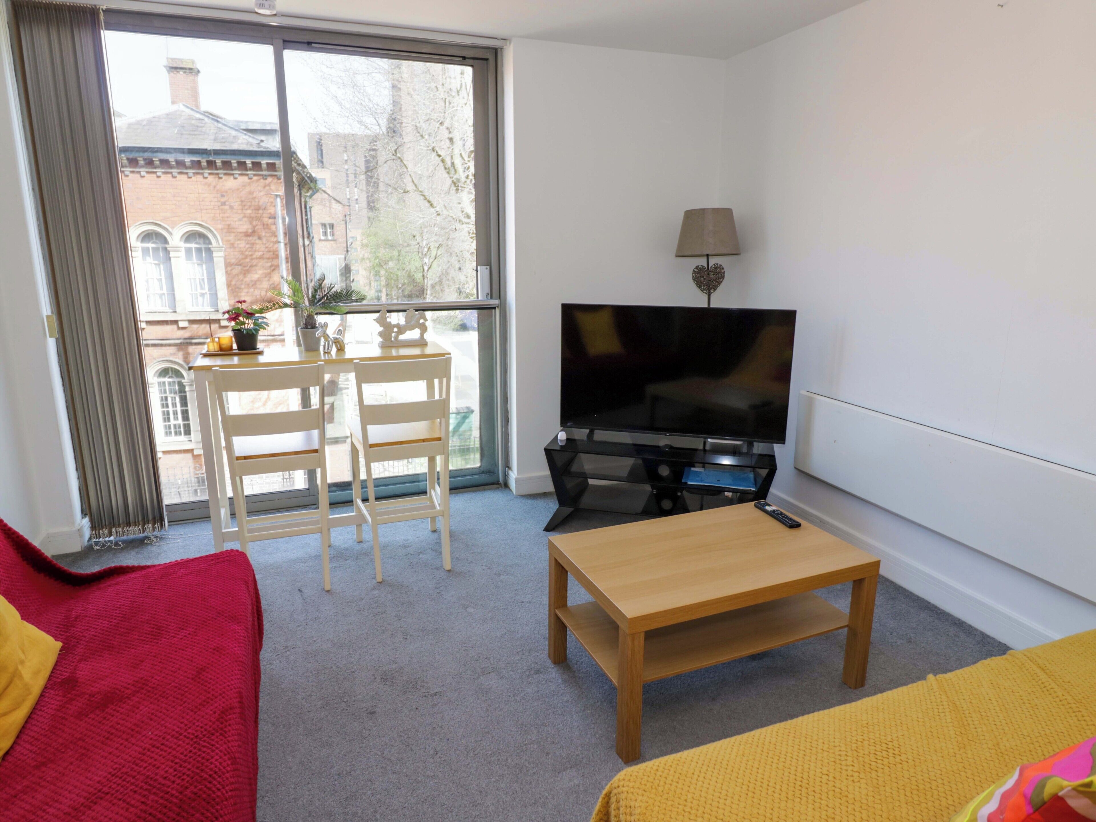 A modern room with a TV, a wooden table, a red sofa, a yellow sofa, and a balcony with a view of buildings and trees.