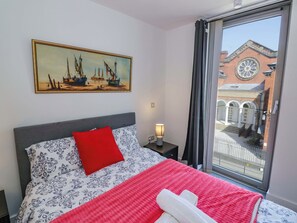 Une chambre d’hôtel avec un lit, un tableau représentant des navires, une fenêtre donnant sur un bâtiment à arcades et un oreiller rouge.