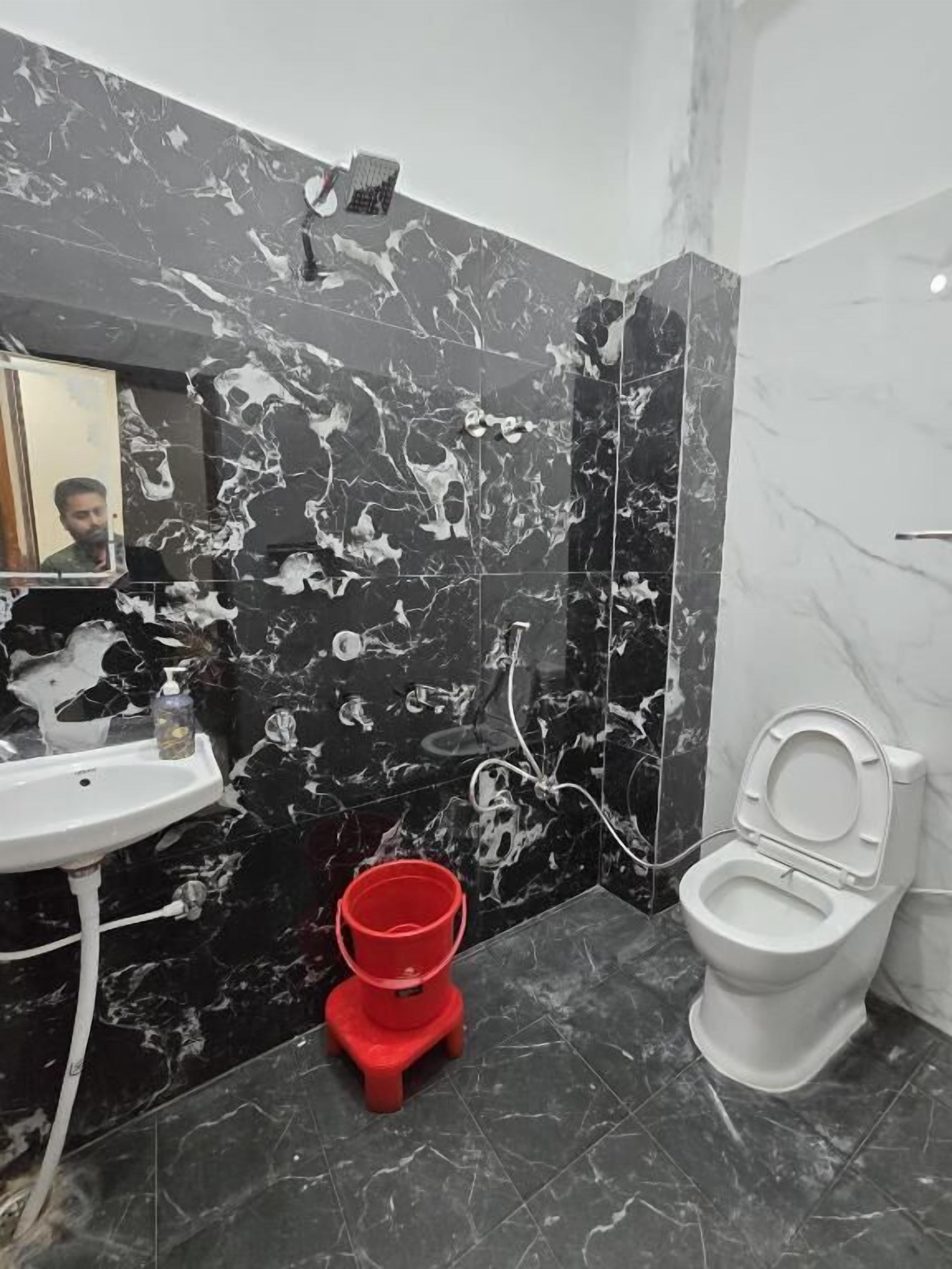 A bathroom with a marble wall, a sink, a toilet, and a red bucket.