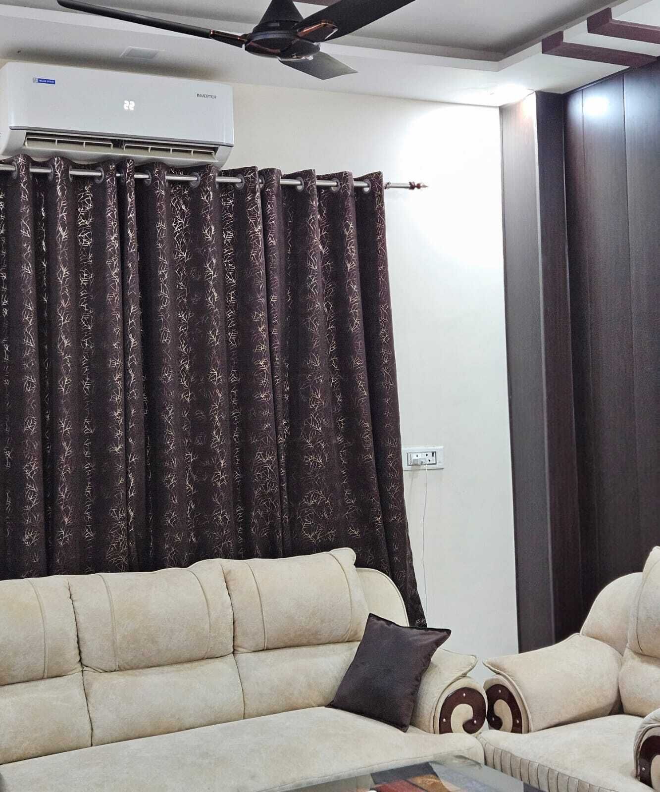 Un salón con sofá color beige, cortina estampada oscura, unidad de aire acondicionado y ventilador de techo.