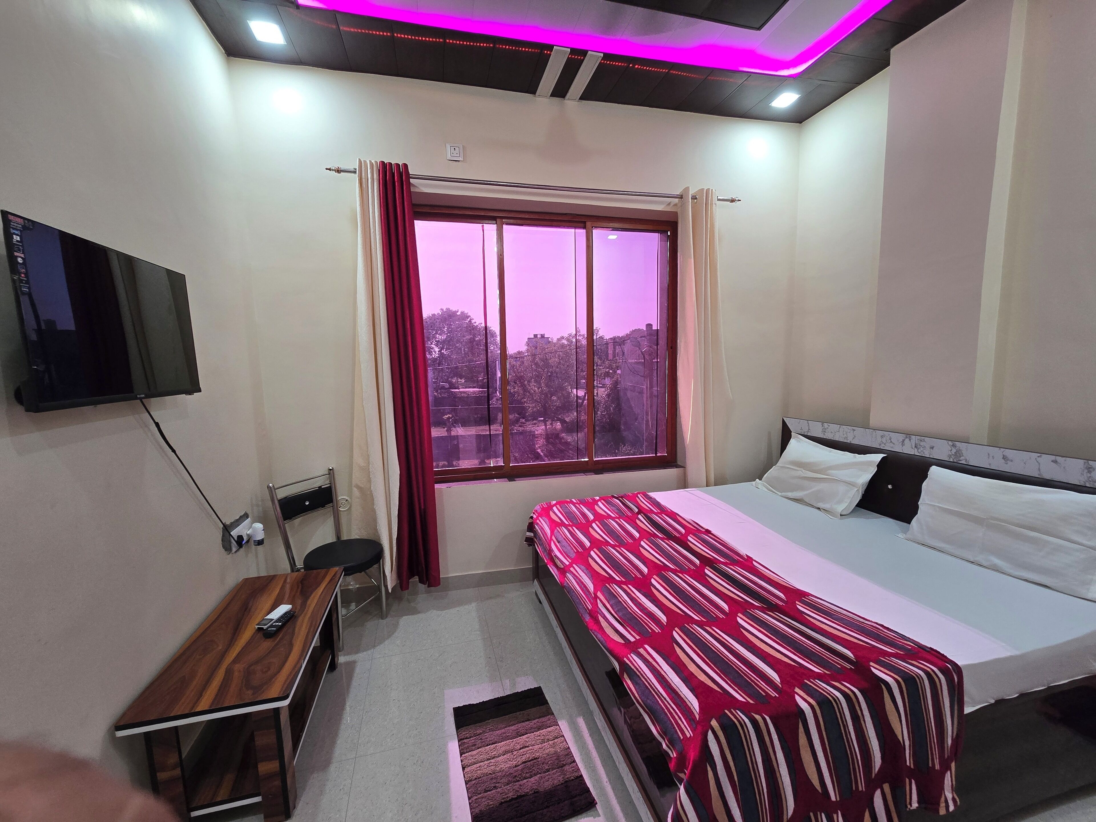Ein Schlafzimmer mit einem Bett, einem Schreibtisch, einem Stuhl, einem an der Wand montierten Fernseher und einem Fenster mit Vorhängen.