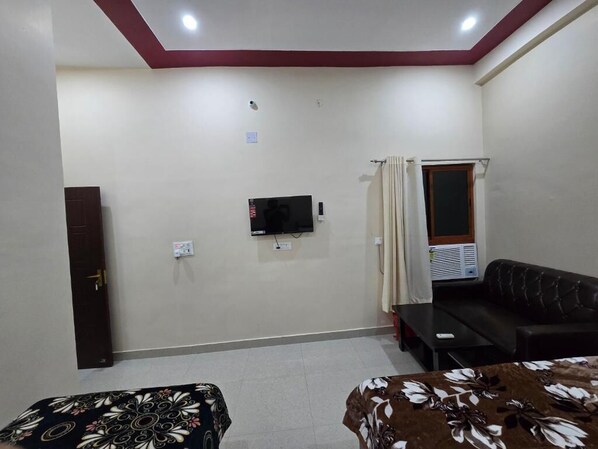 A room with a television mounted on the wall, a sofa, a small table, and a door. - Hotel Siddhi Palace (Ayodhya)