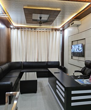 A modern living room with a sectional sofa, a glass coffee table, a desk with a chair, and a mounted TV. - Hotel Siddhi Palace (Ayodhya)