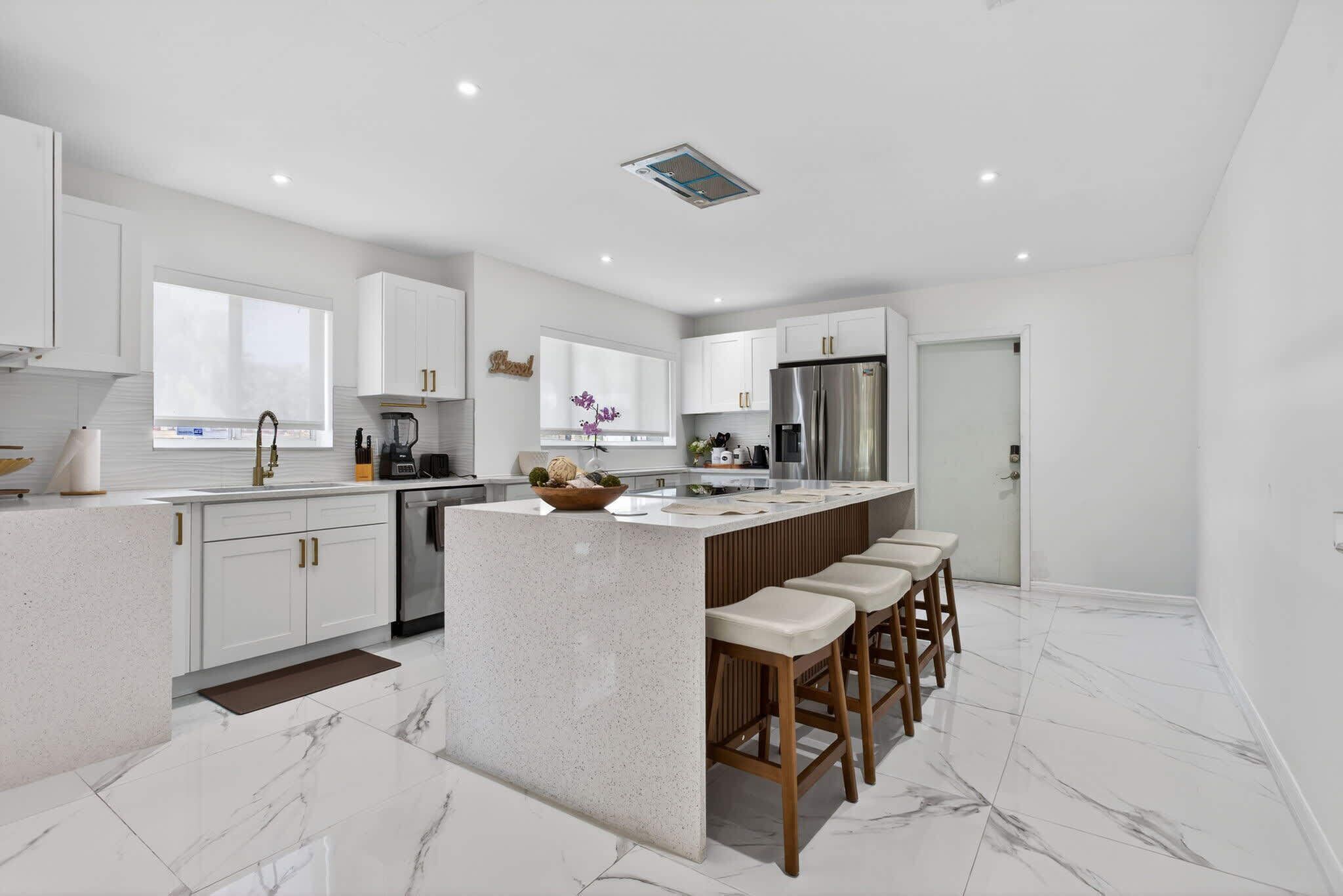 A modern kitchen with white cabinetry, a central island with bar stools, a stainless steel refrigerator, and a window with a view.