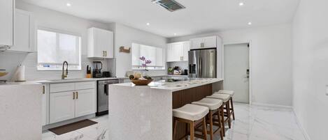 A modern kitchen with white cabinetry, a central island with bar stools, a stainless steel refrigerator, and a window with a view.