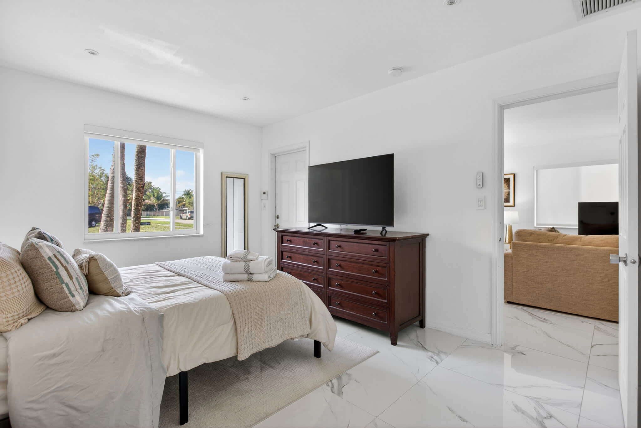 A bedroom with a bed, a dresser, a television, and a view of palm trees outside.