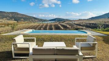 A pool with a view of hills, two white lounge chairs, and a grassy area.