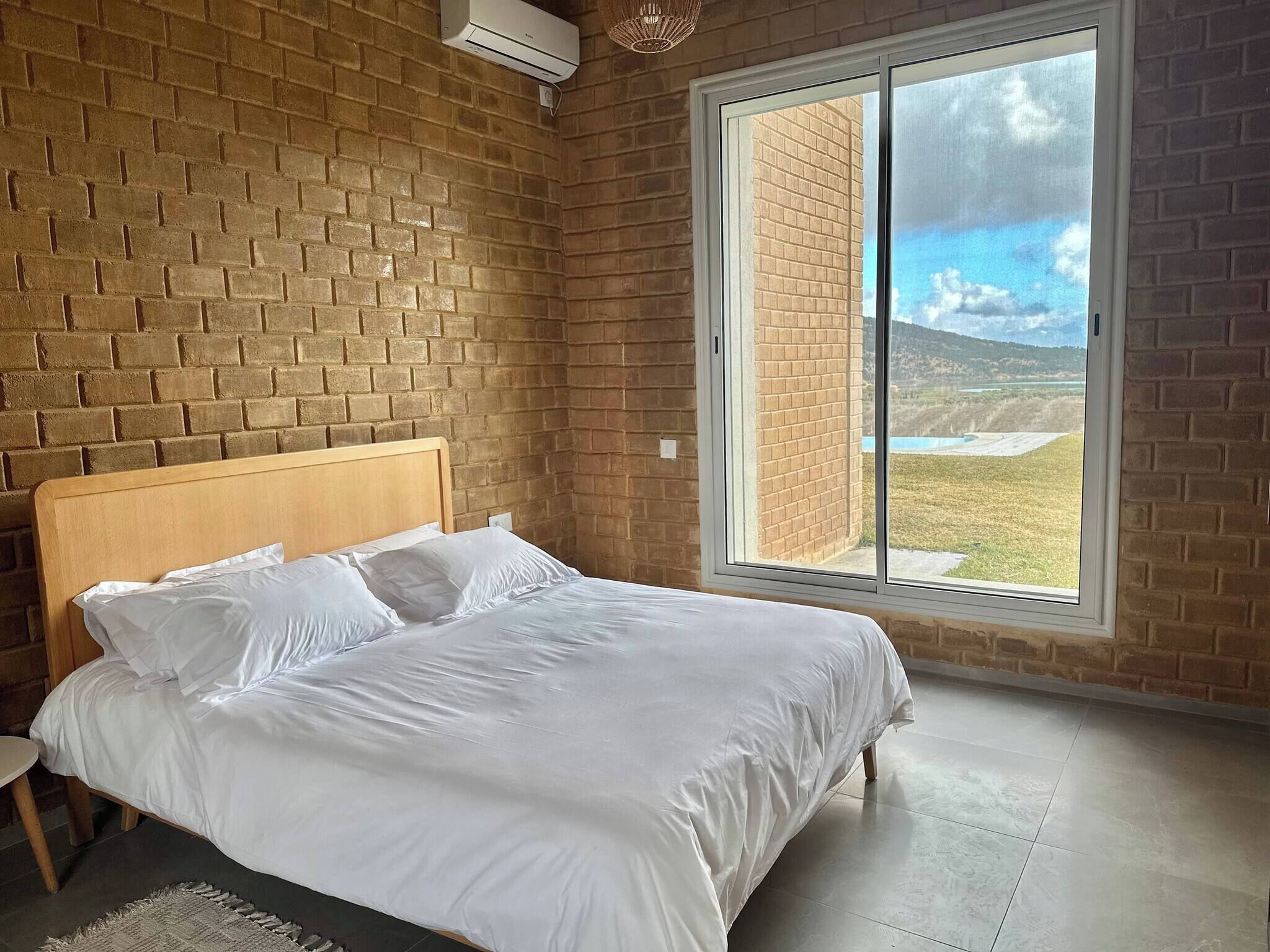 A bedroom with a brick wall, a wooden bed, a large window with a view of a landscape, and an air conditioning unit.