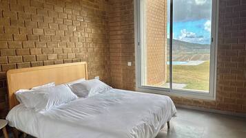 A bedroom with a brick wall, a wooden bed, a large window with a view of a landscape, and an air conditioning unit.