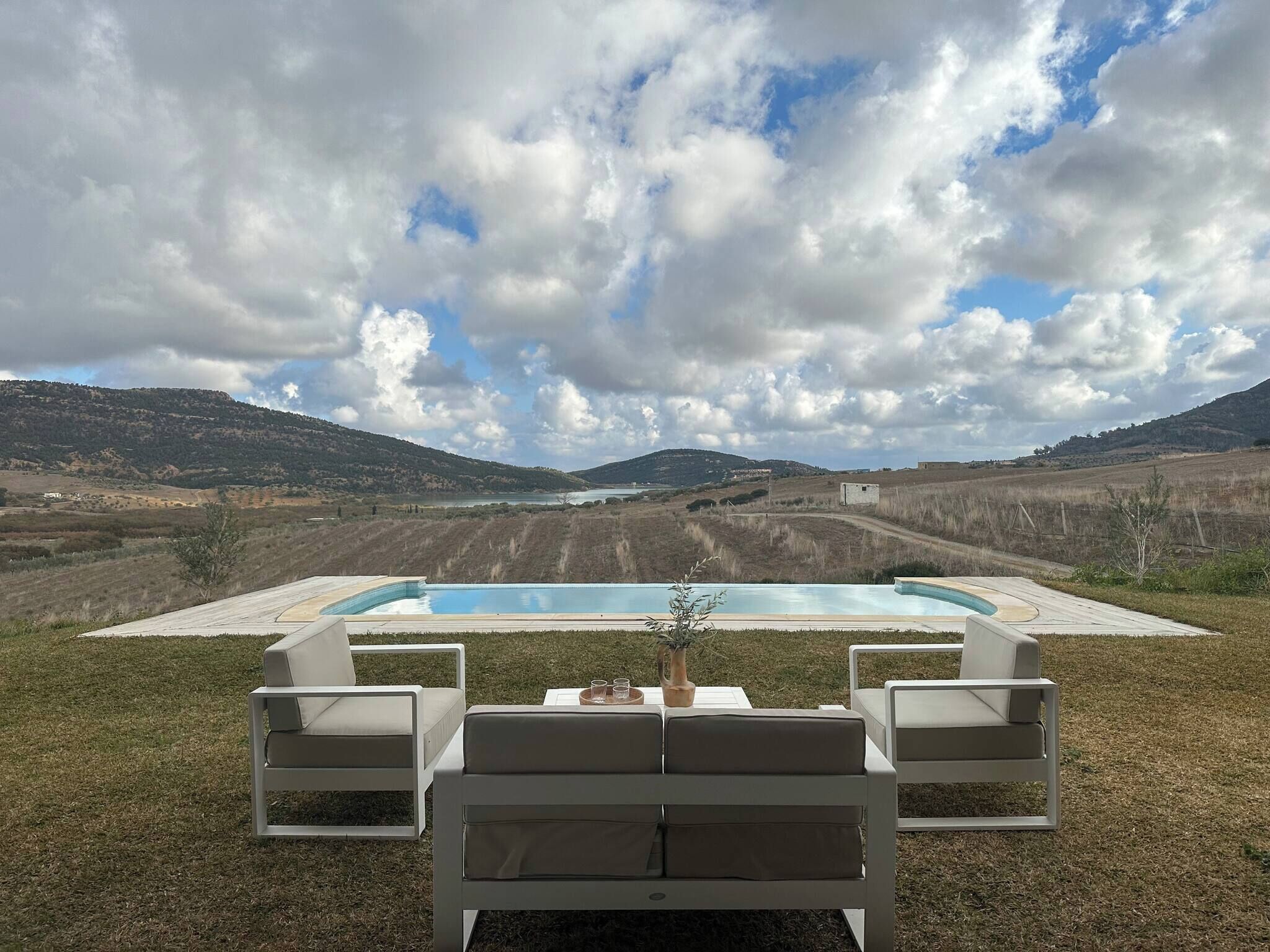 A pool with lounge chairs and a table, surrounded by a grassy area and hills in the background.