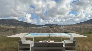 A pool with lounge chairs and a table, surrounded by a grassy area and hills in the background.