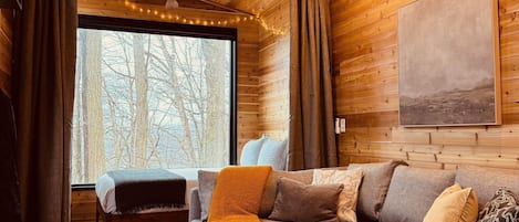 A cabin interior with wooden walls, a sofa, a bed, and a large window with a view of trees.