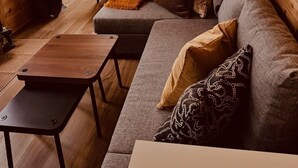 L’intérieur chaleureux d’un chalet, avec un revêtement mural en bois, une grande fenêtre donnant sur les arbres, un canapé agrémenté de coussins et une petite table en bois.