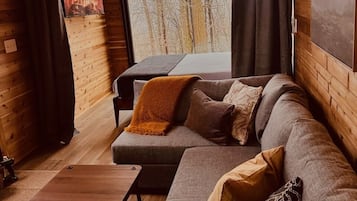 A cozy cabin interior with a wooden interior, a large window with a view of trees, a sofa with cushions, and a small wooden table.