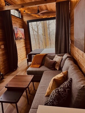 A cozy cabin interior with a wooden interior, a large window with a view of trees, a sofa with cushions, and a small wooden table. - The Oak Tiny House at Cyprus Trail (Rockbridge)