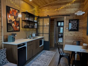 A modern kitchen with wooden cabinetry, a white countertop, and a sink. There is a dining area with a table and chairs, and a large mirror on the wall. - The Oak Tiny House at Cyprus Trail (Rockbridge)