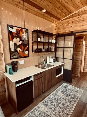 A modern kitchen with wooden cabinetry, a built-in sink, a microwave, and a coffee machine. - The Oak Tiny House at Cyprus Trail (Rockbridge)