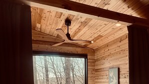 Une pièce aux murs lambrés, avec un lit, un ventilateur de plafond et une grande fenêtre donnant sur une forêt.