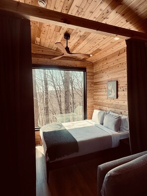 A wooden-paneled room with a bed, a ceiling fan, and a large window overlooking a forest. - The Oak Tiny House at Cyprus Trail (Rockbridge)