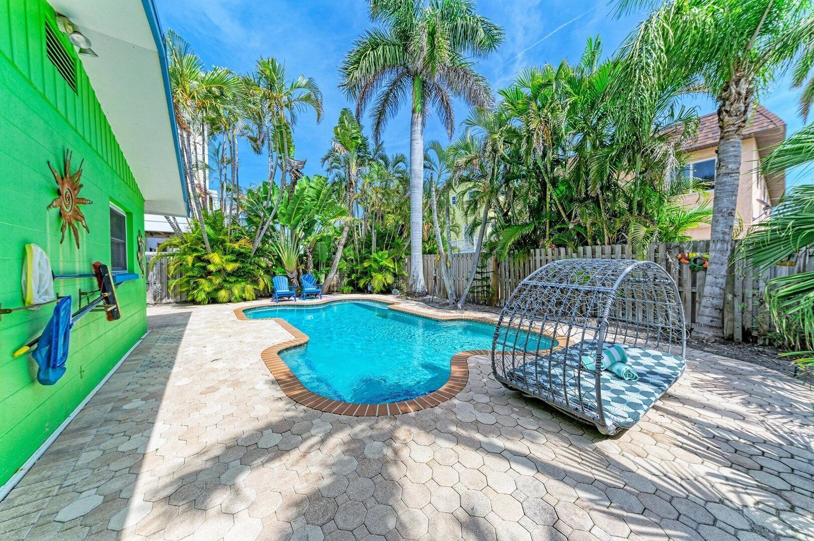 A swimming pool with a hexagonal tile border, surrounded by palm trees and a green building with a sun decoration.