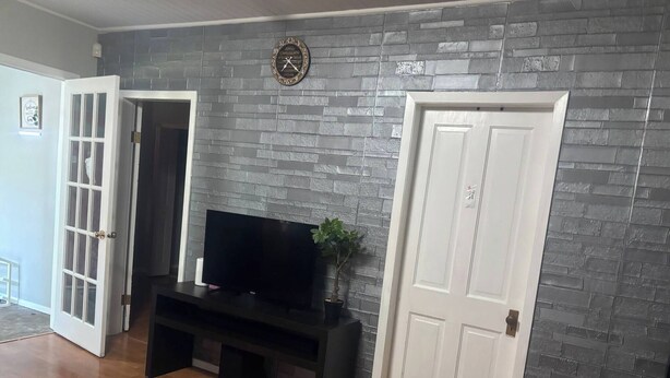 A room with a brick-patterned wall, a black entertainment unit, a white door, and a wooden floor. - Quiet Heritage Bungalow Entire Home Geodome (Edmonton)