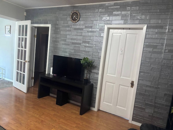 A room with a brick-patterned wall, a black entertainment unit, a white door, and a wooden floor. - Quiet Heritage Bungalow Entire Home Geodome (Edmonton)