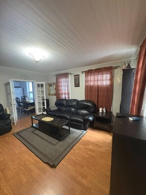 A living room with a black leather sofa, a coffee table, a TV stand, and a door leading to a dining area. - Quiet Heritage Bungalow Entire Home Geodome (Edmonton)