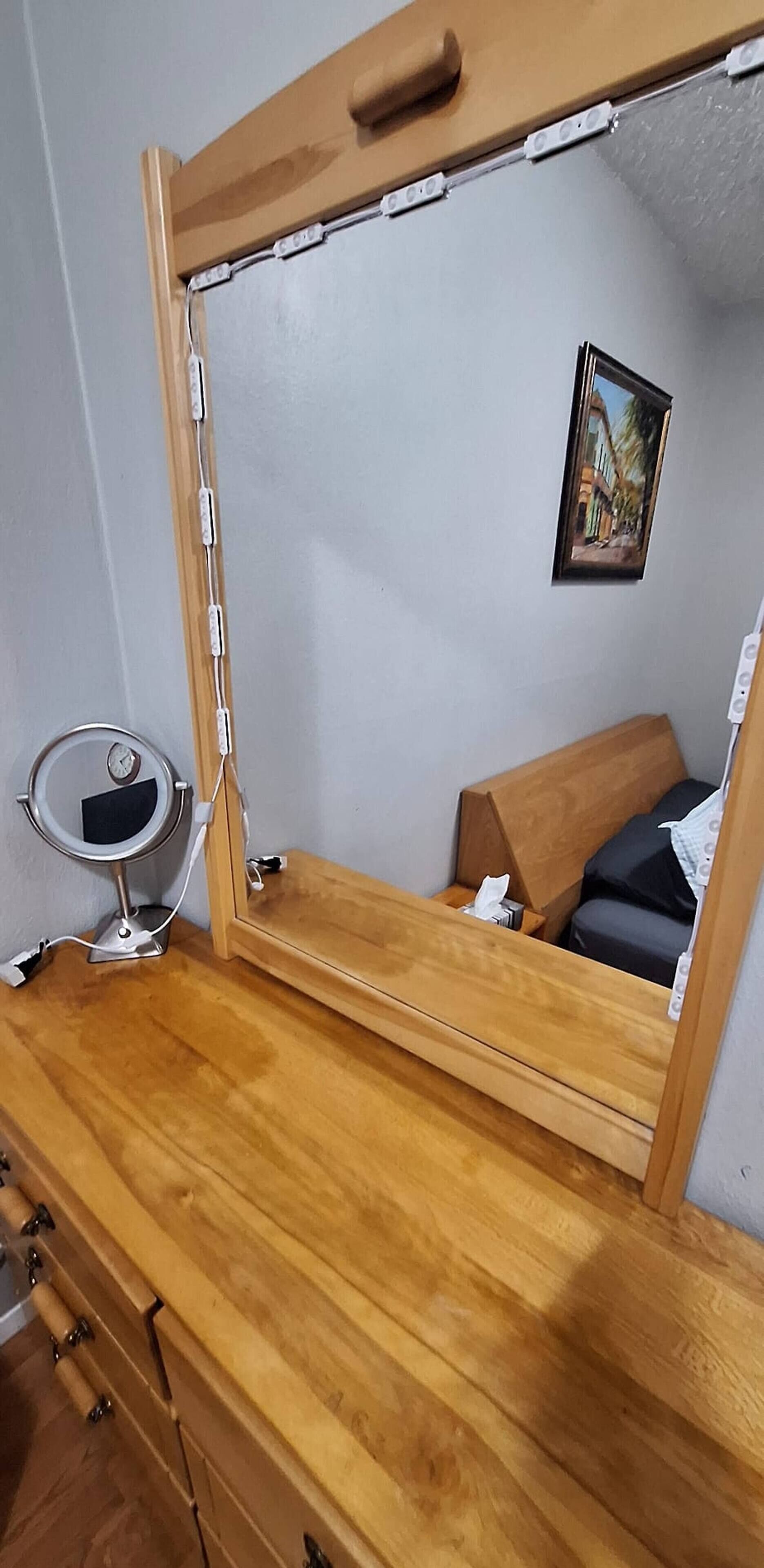 Een houten dressoir met een spiegel, een klein rond tafeltje met een lamp en een ingelijst schilderij aan de muur.