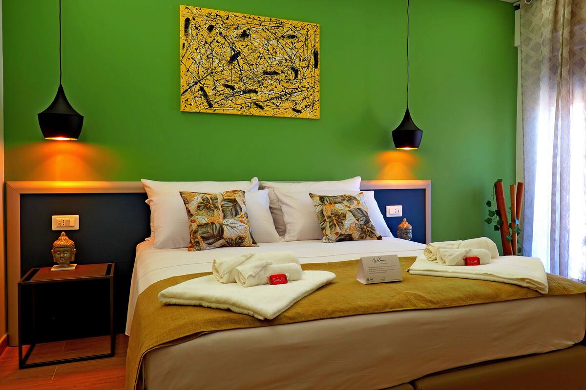 A hotel room with a green wall, a bed with floral pillows, two black pendant lights, and a Buddha statue on the nightstand.