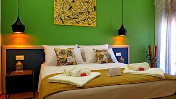 Ein Hotelzimmer mit grüner Wand, einem Bett mit floralen Kissen, zwei schwarzen Pendelleuchten und einer Buddha-Statue auf dem Nachttisch.