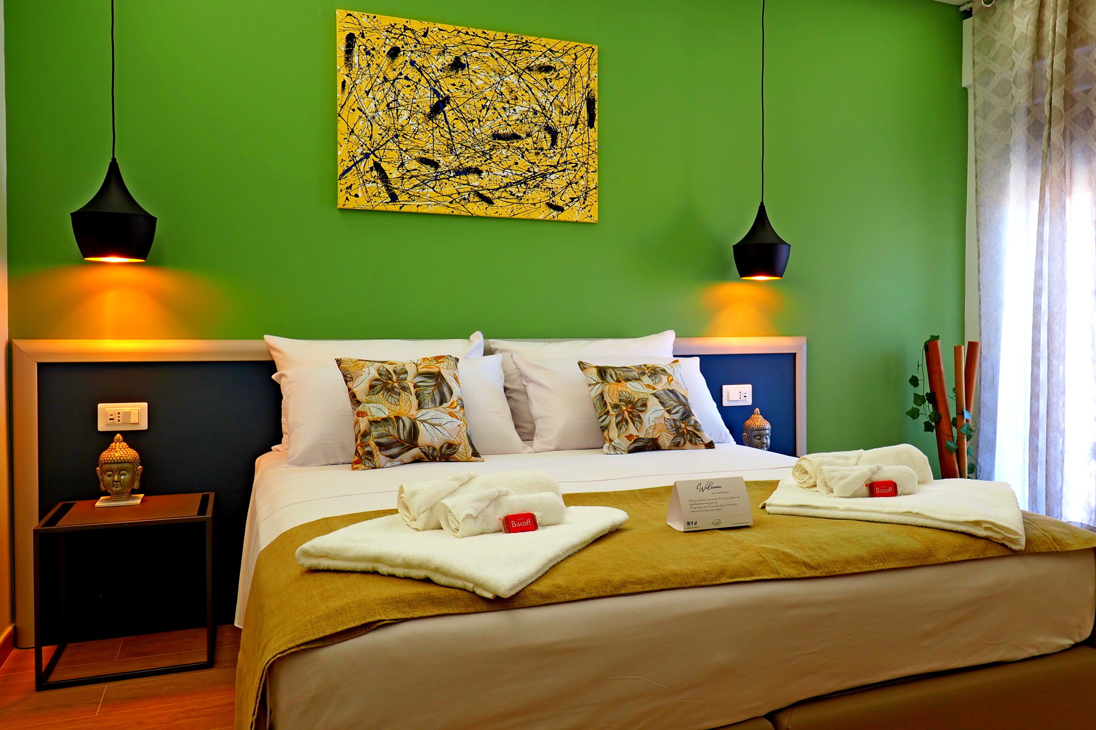 A neatly made bed with a green wall, two black pendant lights, and a Buddha statue on a nightstand.