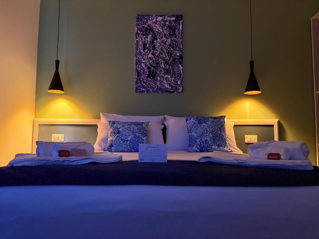 A hotel room with a bed, two bedside lamps, a wall art piece, and a sign on the bed.