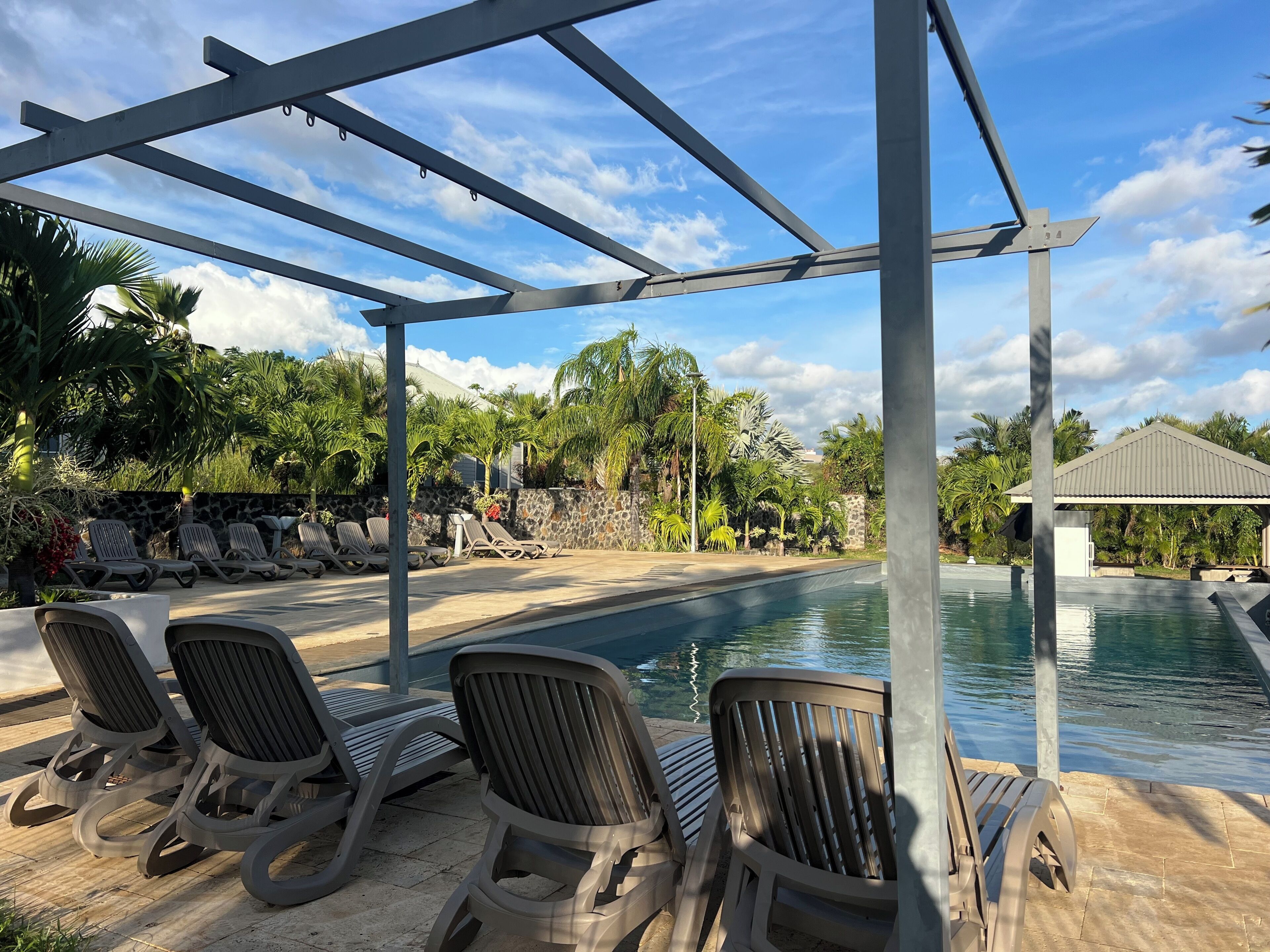A pool area with lounge chairs, a pergola, and a stone wall with tropical plants.