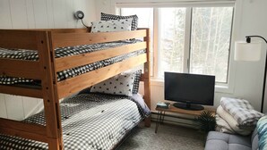 A bunk bed with checkered bedding, a television on a wooden stand, a small table, a sofa, and a window with a view of trees. - Lovely 2-bedroom condo in Bromley Village perfect for a relaxing getaway (Peru)