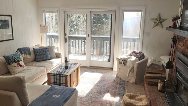 A cozy living room with a sofa, a coffee table, a fireplace, and a balcony with a view of trees. - Lovely 2-bedroom condo in Bromley Village perfect for a relaxing getaway (Peru)