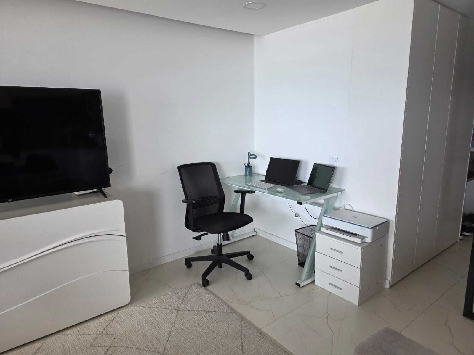 Um escritório moderno com uma televisão de tela plana, uma mesa de vidro com dois laptops, uma cadeira de escritório preta e armários brancos.