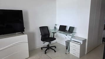Um escritório moderno com uma televisão de tela plana, uma mesa de vidro com dois laptops, uma cadeira de escritório preta e armários brancos.