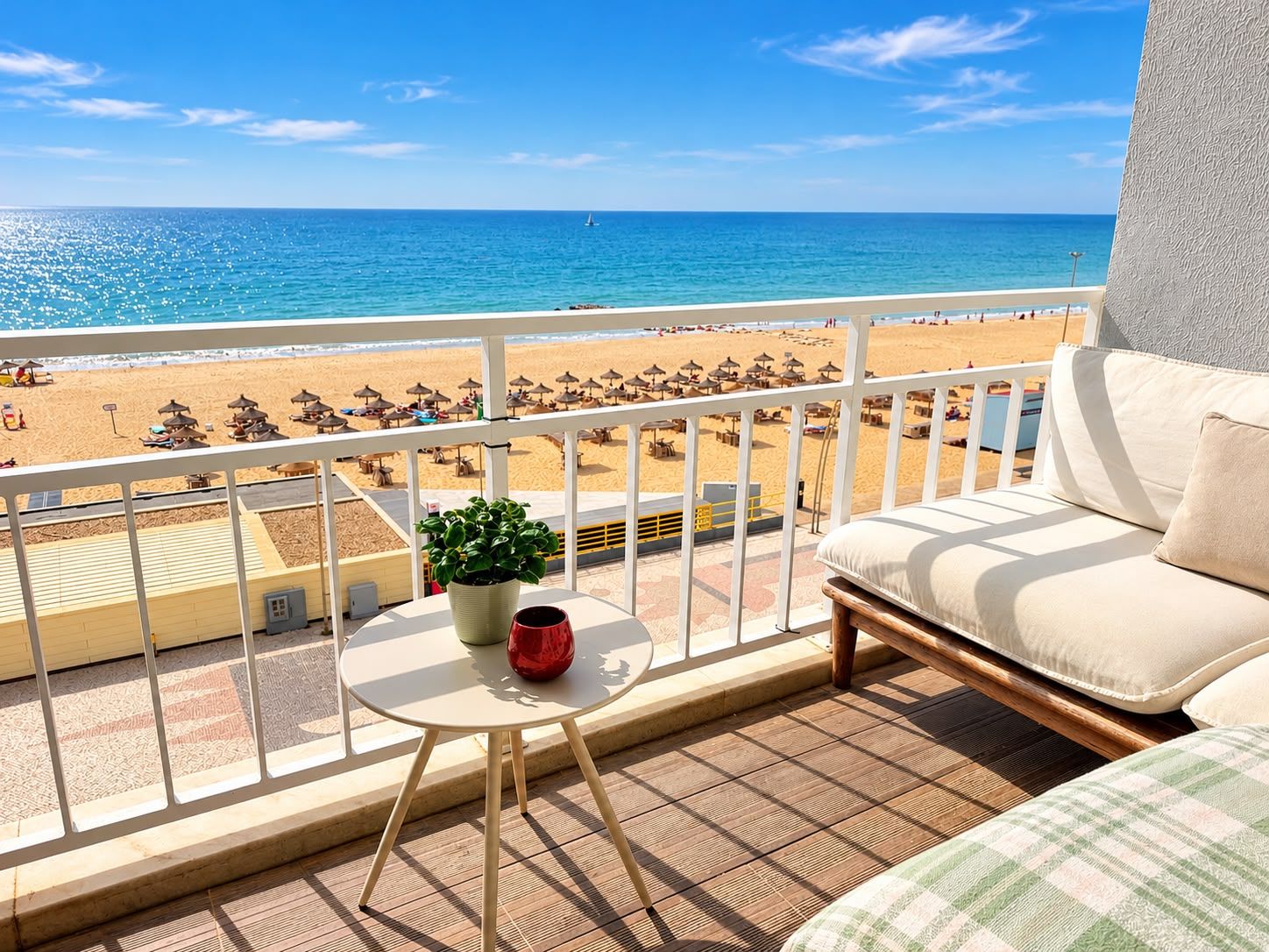Un balcon donnant sur une plage, une table ornée d’une plante et d’un vase rouge, et un canapé avec un coussin.