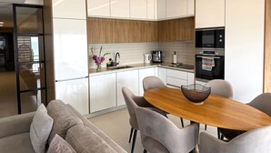 Uma cozinha moderna com armários brancos, bancada de madeira e um forno Bosch. Há uma área de jantar com mesa de madeira e cadeiras cinzas.