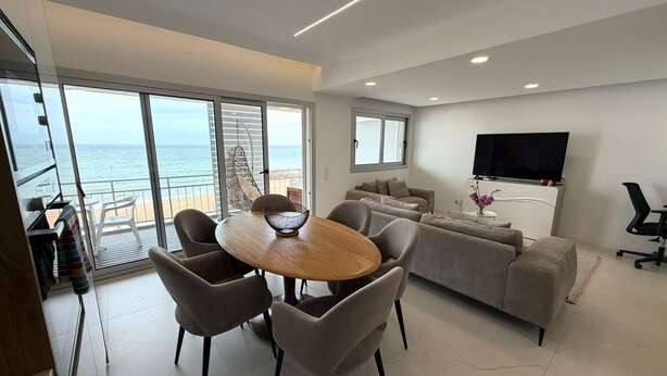 Uma sala de estar moderna com uma grande janela com vista para o mar, uma área de jantar com mesa redonda e uma TV de tela plana fixada na parede.