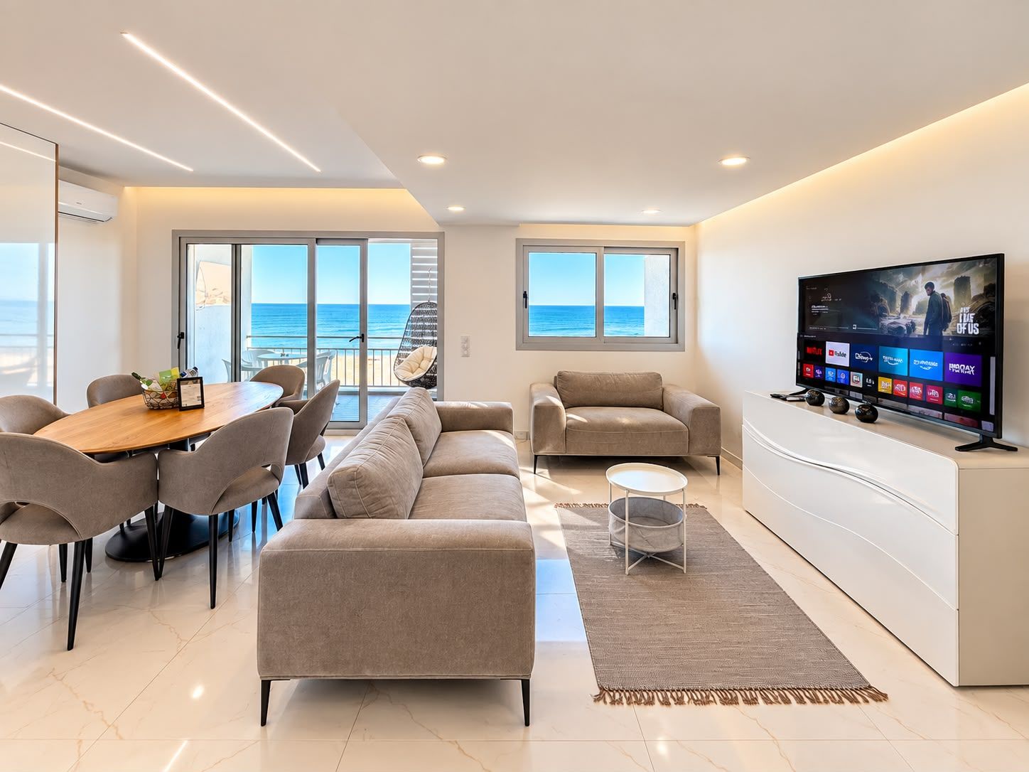 Un salon moderne comprenant un canapé, une table à manger avec des chaises, un téléviseur et une vue sur la mer grâce à de grandes fenêtres.