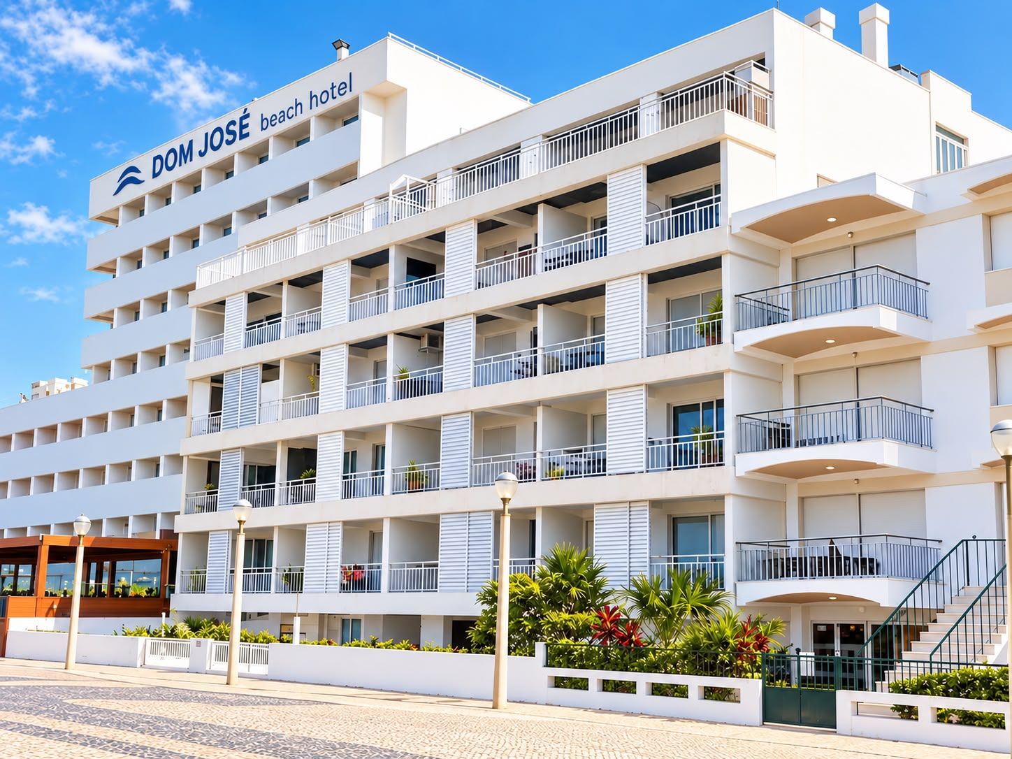 Um hotel de vários andares com varandas, uma placa com a inscrição 'DOM JOSÉ beach hotel' e uma área pavimentada na frente.