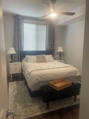 Une chambre à coucher comprenant un lit, des tables de chevet, une commode, un ventilateur de plafond, une fenêtre avec des stores et un tapis à motifs.