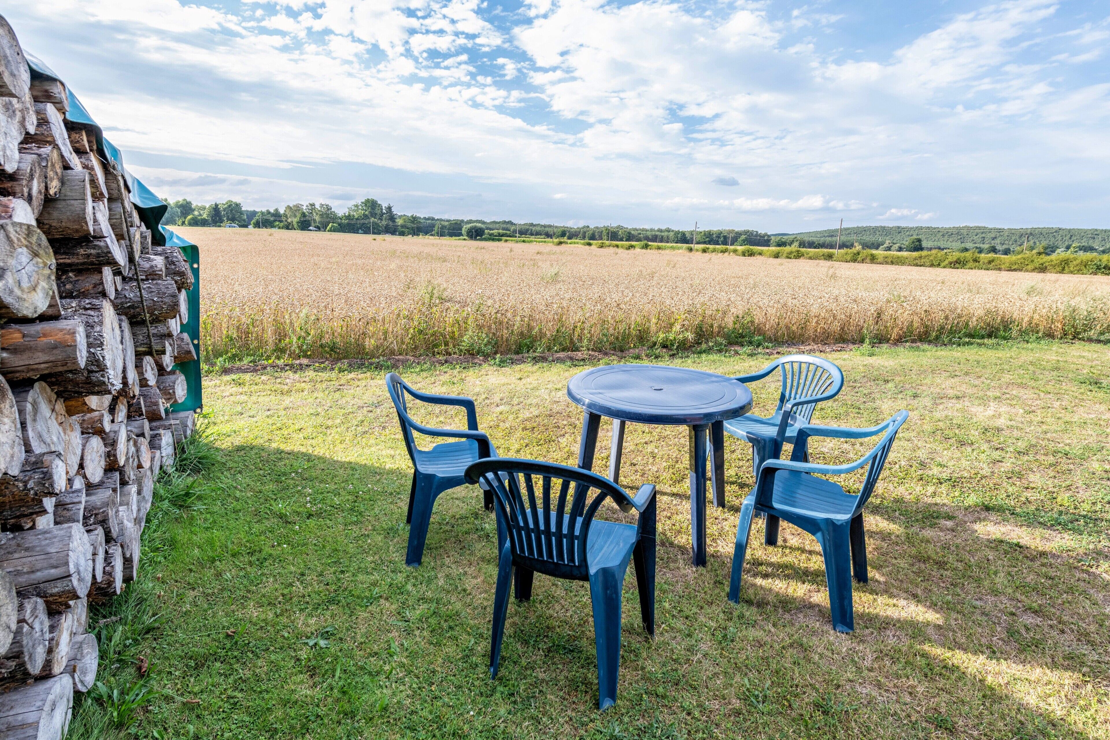 Ein rustikaler Sitzbereich im Freien mit Tisch und Stühlen, einem Holzstoß und einem Feld im Hintergrund.