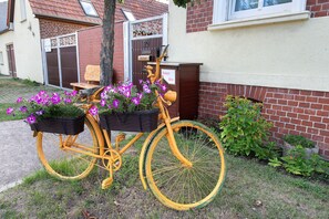 En gul cykel med blomsterkurve, parkeret på en græsplæne foran et hus med en murstensfacade og en postkasse.