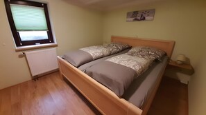Una cama individual con cabecera, una ventana con persianas, un radiador, un cuadro colgado en la pared y una mesita de noche con lámpara.