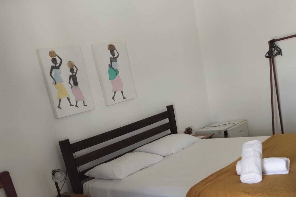 A hotel room with two single beds, a bedside table with a lamp, a wall-mounted heater, and two artwork pieces depicting women.