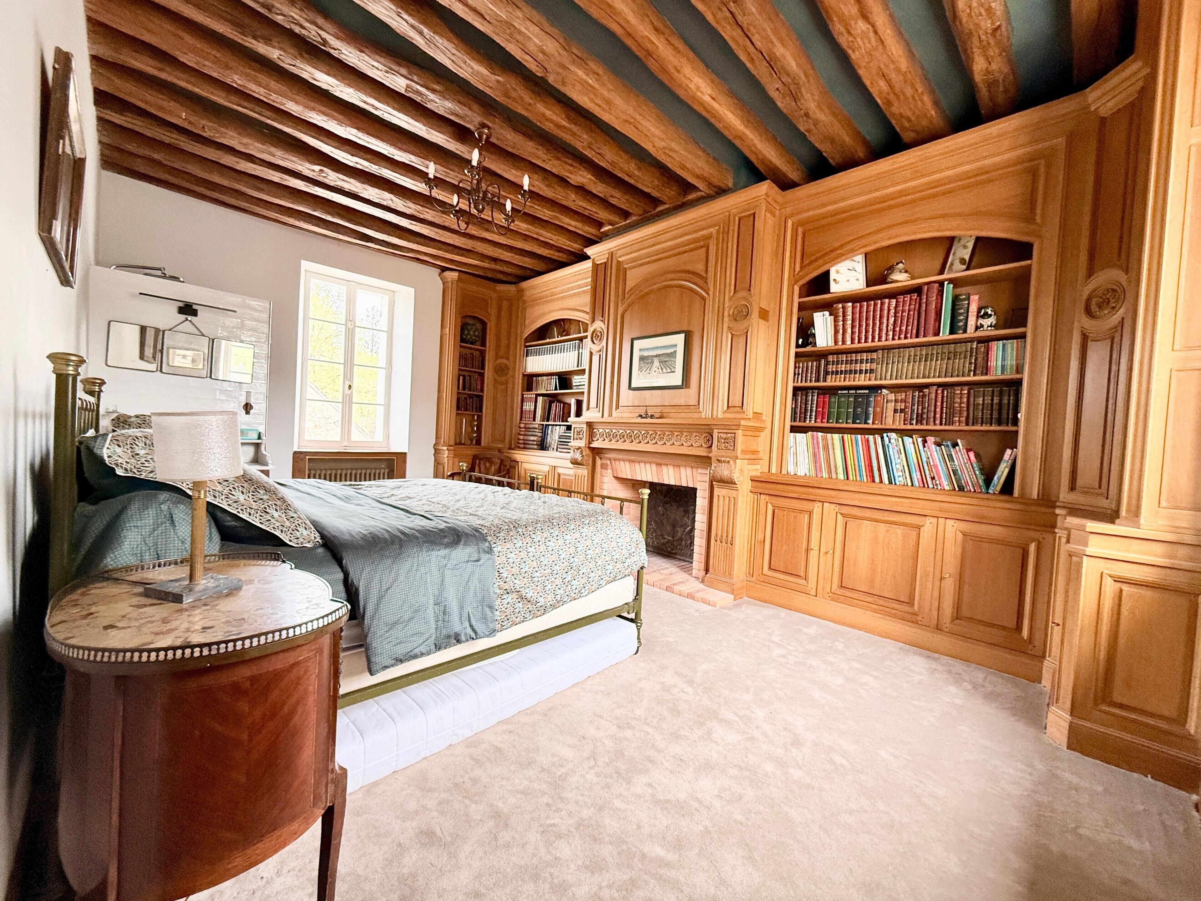 A bedroom with a bed, a bedside lamp, a fireplace, a bookshelf, and a window.