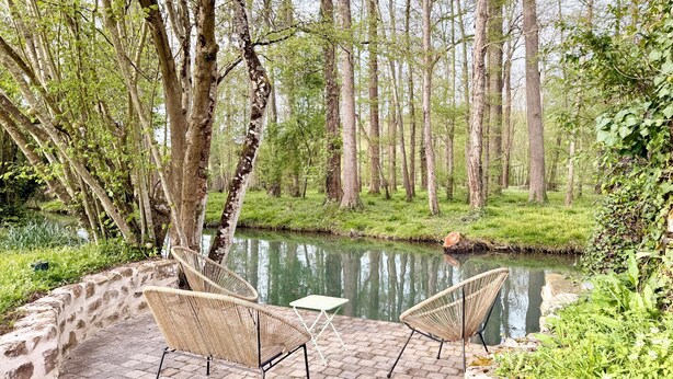 A serene outdoor seating area with wicker chairs and a table, surrounded by lush greenery and a calm water body. - Old Presbytery on the waterfront - pets welcome! (Nanteau-sur-Lunain)