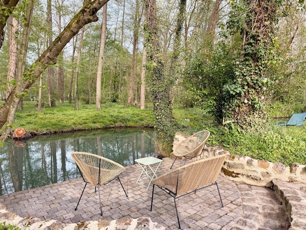 A serene outdoor seating area with wicker chairs and a table, overlooking a calm river and lush greenery.
